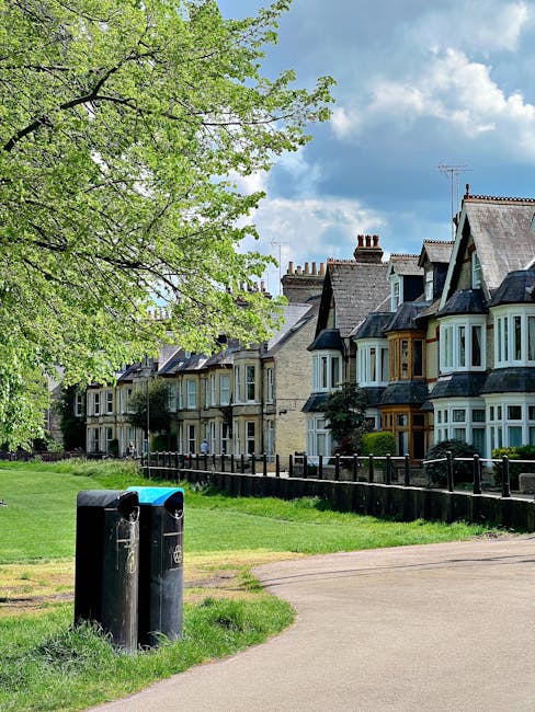 A suburban street scene featuring a row of traditional terraced houses with bay windows, brick facades, and pitched roofs, situated behind a black wooden fence. In the foreground, there are two black 